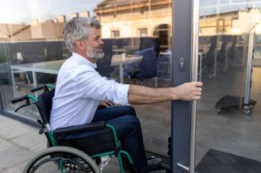 Ofis kapısında. Gri saçlı, sakallı, tekerlekli sandalyedeki olgun adam ofisin kapısını açıyor.