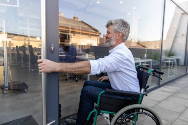 Ofis kapısında. Gri saçlı, sakallı, tekerlekli sandalyedeki olgun adam ofisin kapısını açıyor.