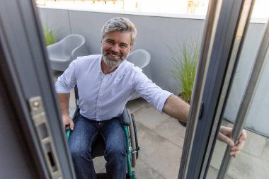 Ofis kapısında. Gri saçlı, sakallı, tekerlekli sandalyedeki olgun adam ofisin kapısını açıyor.