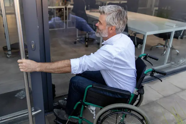 stock image At the office door. Gray-haired bearded mature man on a wheelchair opening the door to the office