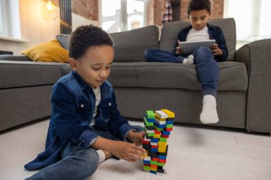 Afro-Amerikalı çocuklar oturma odasında tablet ve inşaatçıyla oynuyorlar.