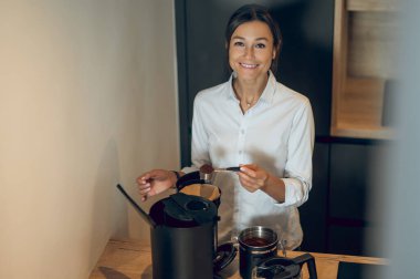 Kahve yapıyorum. Koyu saçlı genç bayan mutfakta kahve makinesinde kahve yapıyor.