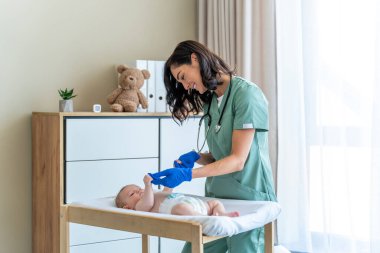 Mavi eldivenli gülümseyen kadın çocuk doktoru klinikte küçük parmağını tutan bebeği muayene ediyor.