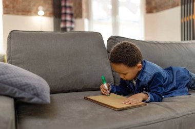 Neşeli çocuk elinde pastel boya kalemi ile kanepede boş vakitlerde kağıda çizimler çiziyor.