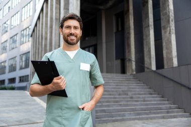 Sakallı profesyonel bir doktor elinde tıbbi evraklarla modern kliniğin yakınlarında poz veriyor.