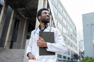 Sakallı çok kültürlü bir doktor elinde tıbbi belgelerle modern kliniğin yakınlarında poz veren bir pano tutuyor.