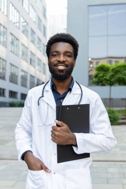 Sakallı çok kültürlü bir doktor elinde tıbbi belgelerle modern kliniğin yakınlarında poz veren bir pano tutuyor.