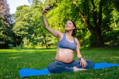 Neşeli hamile kadın yaz yoga eğitimi sırasında parkta selfie çekiyor.