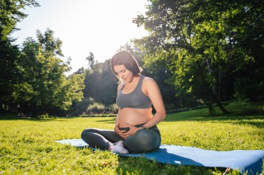 Hamile kadın parkta egzersiz yapıyor. Sıcak yaz güneşinin tadını çıkarıyor. Yeşil çimlerde yoga yapıyor.