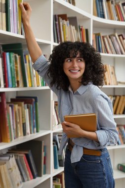 Kütüphane. Kitaplıktaki sevimli, genç, kıvırcık saçlı kız.