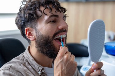 Man with metal braces carefully cleaning his teeth using interdental brush focusing on removing debris between brackets to maintain proper dental hygiene during orthodontic treatment clipart