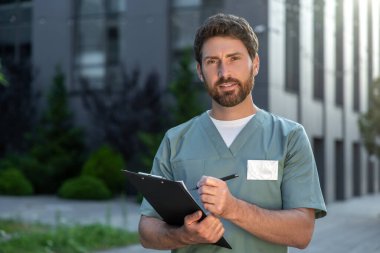 Tıbbi üniforma ve steteskop giyen sakallı bir adam hastanenin dışında hastalarla ilgilenmeye hazır bekliyor.