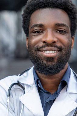 Tıbbi üniforma giyen ve steteskop takan kendine güvenen sakallı bir doktor kliniğin önünde dikiliyor.