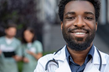 Tıbbi üniforma ve steteskop giyen sakallı bir adam hastanenin dışında hastalarla ilgilenmeye hazır bekliyor.