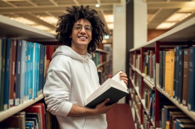 Kütüphanede. Koyu saçlı genç adam kitaplığın yanında duruyor ve kitap okuyor.