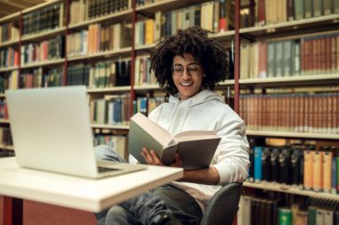 Çalışıyorum. Kıvırcık saçlı genç adam kütüphanede çalışıyor ve ilgili görünüyor.