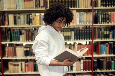 Kitap kurdu. Kütüphanede gözlüklü kıvırcık saçlı genç adam.