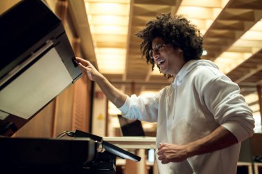 Kopyalıyorum. Kıvırcık saçlı genç adam kütüphanede fotokopi çekiyor.