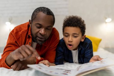 Birlikte kitap okuyoruz. Esmer tenli bir adam, oğluyla birlikte barakadaki resimlere bakıyor.