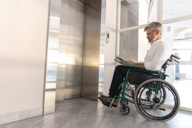 Tekerlekli sandalyedeki sakallı işadamı şirkette asansörü beklerken belgeleri inceliyor.