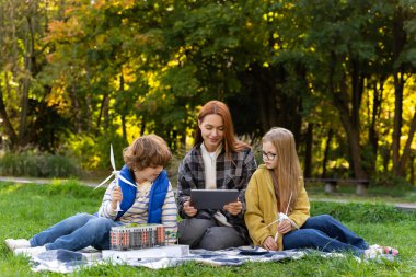 Şehir parkında çocuklarla dijital tablet kullanan genç bayan öğretmen rüzgar türbinlerini, yenilenebilir enerjiyi ve sürdürülebilir kaynakların önemini öğreniyor