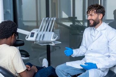 Diş kontrolü sırasında dişçi koltuğunda oturan Afrikalı Amerikalı sakallı bir adam doktoruyla konuşuyor.