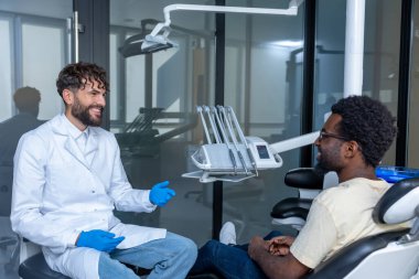 Diş kontrolü sırasında dişçi koltuğunda oturan Afrikalı Amerikalı sakallı bir adam doktoruyla konuşuyor.