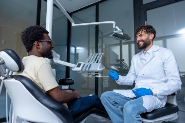 Diş kontrolü sırasında dişçi koltuğunda oturan Afrikalı Amerikalı sakallı bir adam doktoruyla konuşuyor.