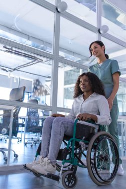 Hastanedeydim. Koyu saçlı hemşire kadın bir hastayla tekerlekli sandalye taşıyor.