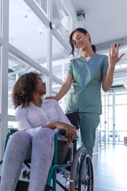 Hastanedeydim. Koyu saçlı hemşire kadın bir hastayla tekerlekli sandalye taşıyor.