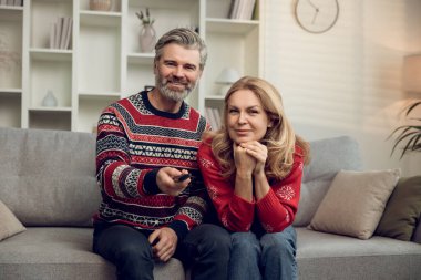 Evde huzurlu bir zaman. Gri saçlı, olgun, yakışıklı bir adam, karısıyla kanepede otururken televizyonu değiştiriyor.