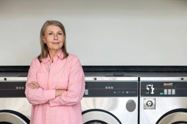 Çamaşırhanede. Çamaşırhanede çamaşır makinesinin yanında duran pembe tişörtlü kendinden emin olgun bir kadın.