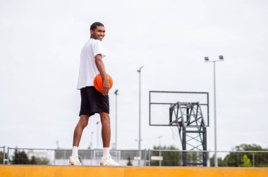 Basketbol mu? Basketbol sahasının yanında elinde topla duran spor kıyafetli uzun boylu bir adam.