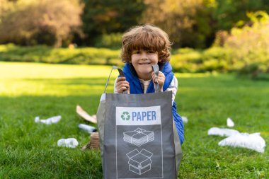 Küçük çocuk, plastiğin geri dönüşümü için sosyal projeye katılarak parkta kağıt çöplerini topluyor.