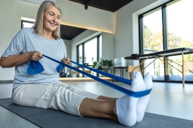 Kafkasyalı olgun bir kadın rehabilitasyon merkezinde geçirdiği travma sonrası bacağını esnetmek için direnç bandı kullanıyor.