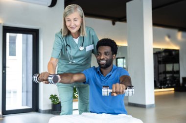 Physical therapy. African american young man working on his arms , female doctor guiding him clipart