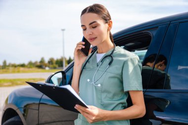 Phone call with a patient. Female doctor standing near the car and talking on the phone clipart