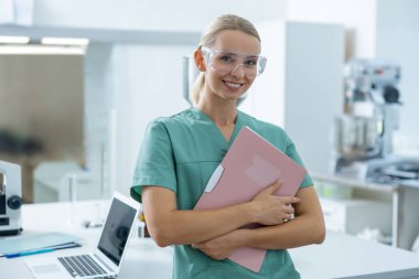 Laboratuvar önlüklü ve koruyucu gözlüklü genç kadın portresi hastane laboratuvarında elinde kağıt klasörle çalışıyor.