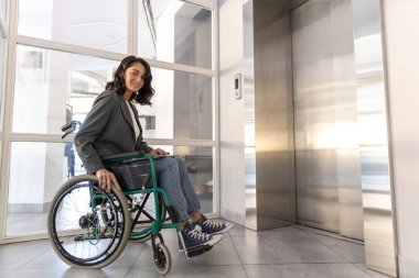 Tekerlekli sandalyedeki iş kadını ofisinde çalışıyor ve yukarı çıkmak için asansörü kullanıyor.