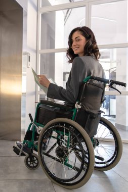 Tekerlekli sandalyedeki genç iş kadını ofis binasında asansör beklerken belgeleri inceliyor.