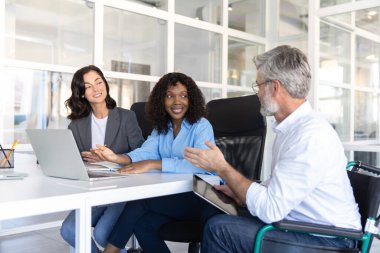 Tekerlekli sandalyedeki işadamı da dahil olmak üzere bir grup işçi laptopun etrafında iş birliği yaparak toplu iş görevlerini çözüyor.