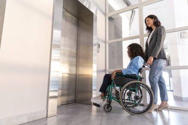 Tekerlekli sandalyedeki engelli genç kadın, kadın meslektaşıyla birlikte asansörde bekliyor.
