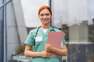 Steteskoplu kızıl saçlı kadın doktor hastane koridorunda poz verirken gülümseyerek üniforma giyiyor.