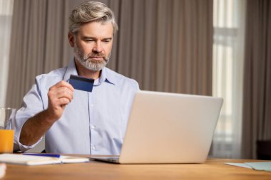 Çevrimiçi ödeme. Gri saçlı, orta yaşlı, sakallı adam masada oturuyor ve internetten ödeme yapıyor.