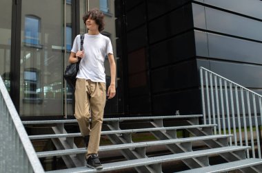 Öğrenci. Beyaz tişörtlü uzun boylu beyaz adam binanın girişindeki merdivenlerde.