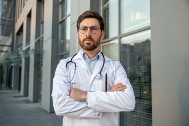 Kendine güvenen bir doktor. Gözlüklü sakallı erkek doktor kararlı görünüyor ve kolları bağlı duruyor.