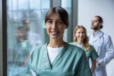 Hastane koridorunda duran üniformalı güzel bayan doktor ve arka planda tıbbi personel.