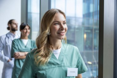 Hastane koridorunda duran üniformalı güzel bayan doktor ve arka planda tıbbi personel.