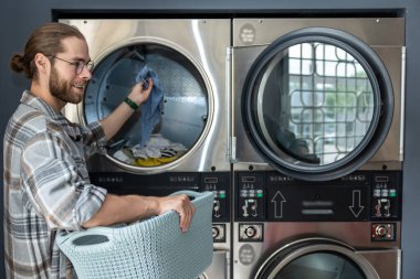 Çamaşırhanedeki sakallı adam çamaşırlarını yıkarken ticari çamaşır makinesi kullanıyor.