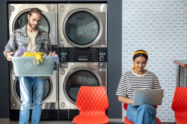 Çamaşırhanedeki kadın dizüstü bilgisayar kullanırken, erkek çamaşır makinesini çamaşır yıkamak için kullanıyor.
